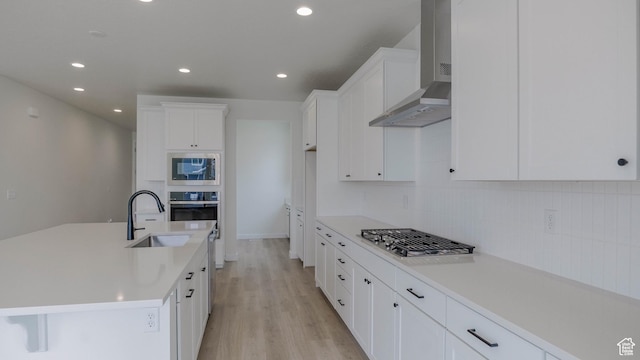 kitchen featuring wall chimney range hood, white cabinetry, light wood-style floors, stainless steel appliances, and a sink