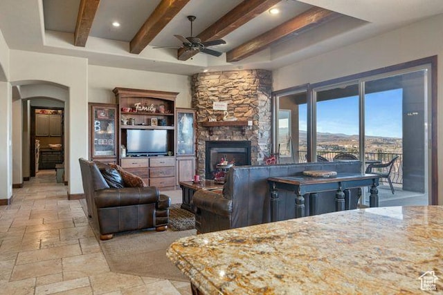 living room with arched walkways, a stone fireplace, baseboards, beam ceiling, and stone tile flooring