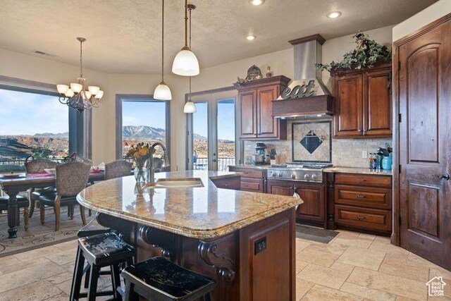 kitchen featuring decorative light fixtures, stone tile flooring, a kitchen island with sink, a sink, and wall chimney range hood
