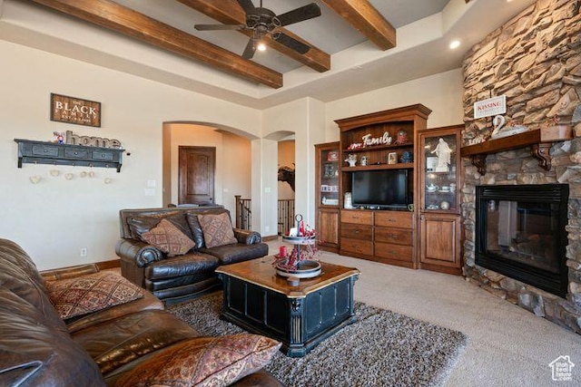 carpeted living room with arched walkways, a stone fireplace, a ceiling fan, baseboards, and beamed ceiling