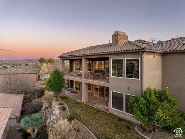 back of house at dusk featuring a patio, a balcony, a tile roof, a chimney, and stucco siding