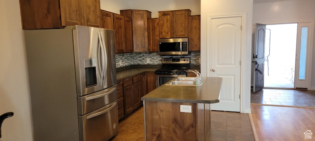 kitchen featuring dark countertops, backsplash, a center island with sink, stainless steel appliances, and a sink