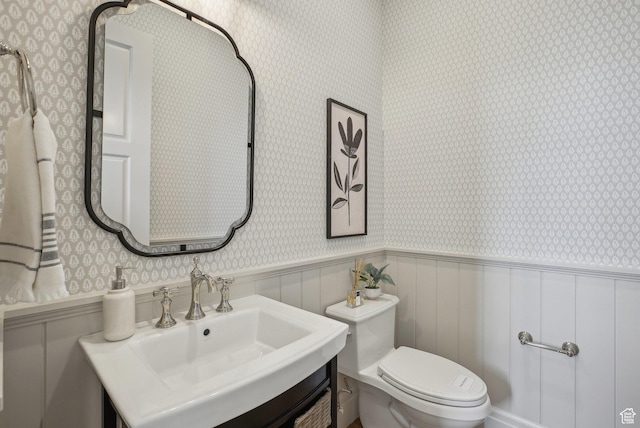 bathroom with a sink, wallpapered walls, toilet, and a wainscoted wall