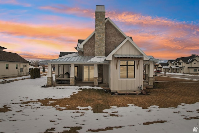 snow covered house with a chimney, covered porch, board and batten siding, a standing seam roof, and metal roof