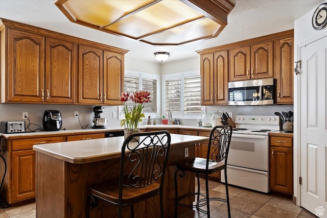 kitchen featuring stainless steel microwave, white electric range, a breakfast bar, light countertops, and brown cabinetry