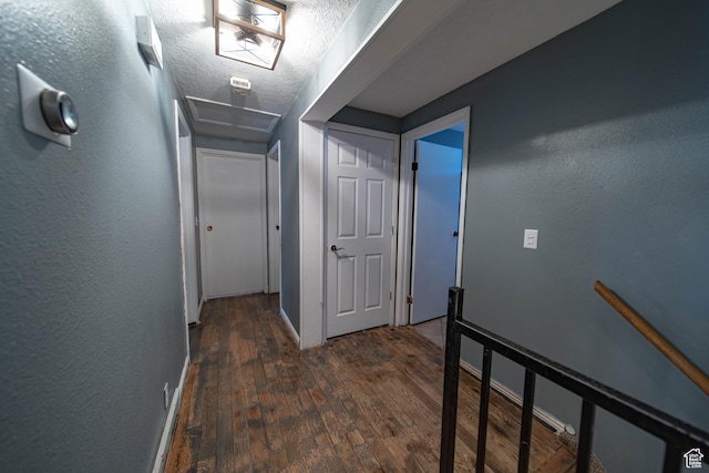 hallway featuring a textured ceiling, a textured wall, dark wood-type flooring, an upstairs landing, and attic access
