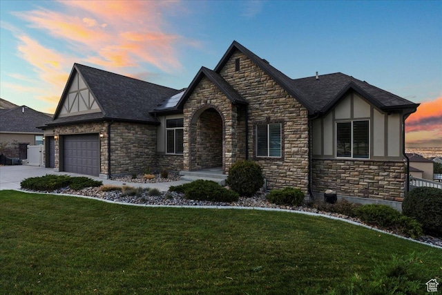tudor-style house with driveway, a shingled roof, an attached garage, a front lawn, and stucco siding