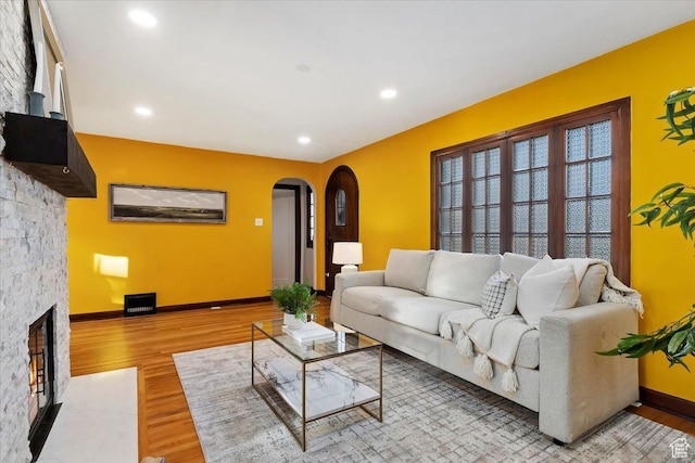 living room featuring arched walkways, a stone fireplace, recessed lighting, baseboards, and light wood-type flooring