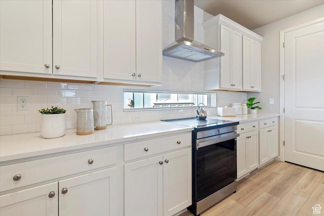 kitchen with stainless steel electric stove, backsplash, white cabinets, light wood-type flooring, and wall chimney exhaust hood