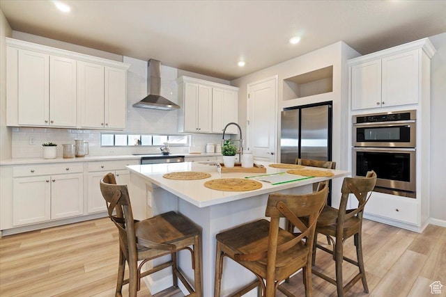 kitchen with light wood finished floors, a kitchen bar, light countertops, appliances with stainless steel finishes, and wall chimney exhaust hood