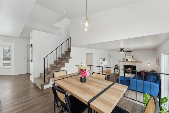 dining space with a ceiling fan, wood finished floors, a warm lit fireplace, baseboards, and stairs