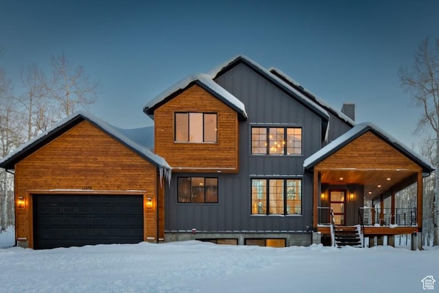 snow covered property with board and batten siding, covered porch, a chimney, and an attached garage