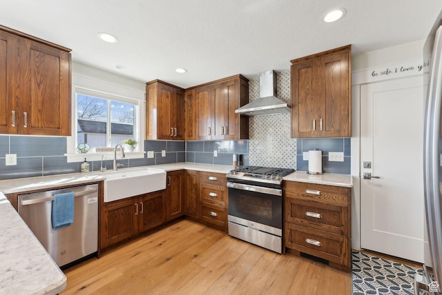 kitchen with a sink, light countertops, wall chimney range hood, appliances with stainless steel finishes, and light wood finished floors