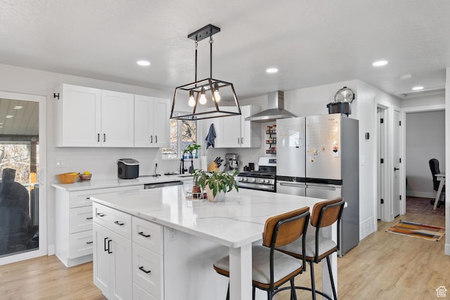 kitchen featuring light wood-style flooring, stainless steel appliances, white cabinetry, wall chimney range hood, and light stone countertops