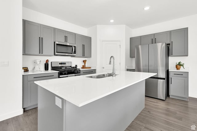 kitchen with gray cabinets, stainless steel appliances, a center island with sink, a sink, and light wood finished floors