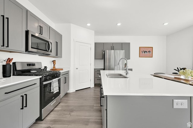 kitchen with stainless steel appliances, an island with sink, a sink, light wood-type flooring, and gray cabinetry