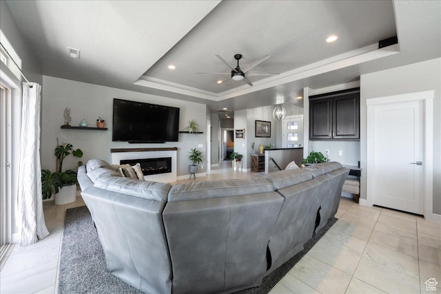 living room featuring visible vents, a ceiling fan, a glass covered fireplace, a tray ceiling, and recessed lighting
