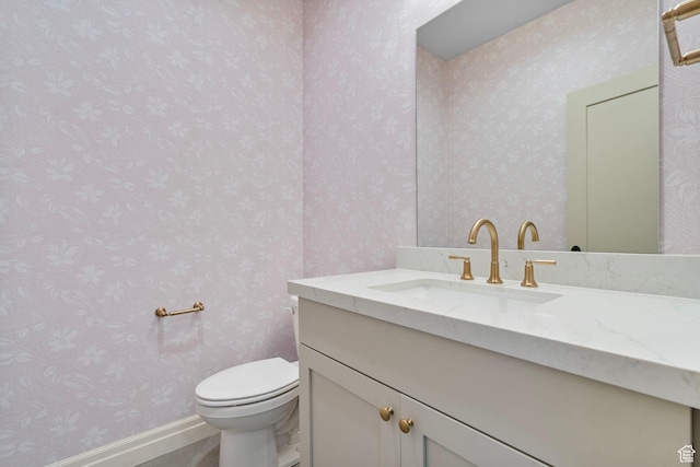 half bathroom with baseboards, vanity, toilet, and wallpapered walls