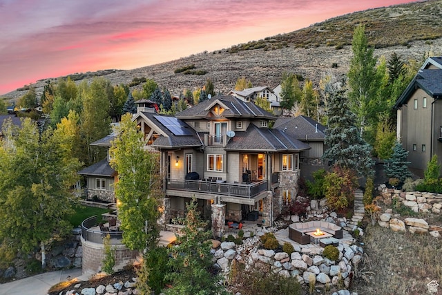 back of house at dusk featuring a balcony, solar panels, a fire pit, stone siding, and a patio area