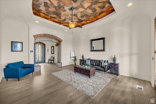 living room with arched walkways, a raised ceiling, visible vents, a ceiling fan, and wood finished floors