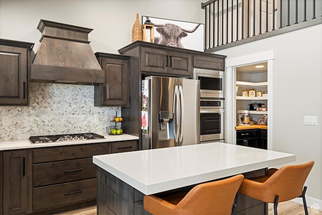 kitchen featuring dark brown cabinetry, a breakfast bar, decorative backsplash, appliances with stainless steel finishes, and custom exhaust hood