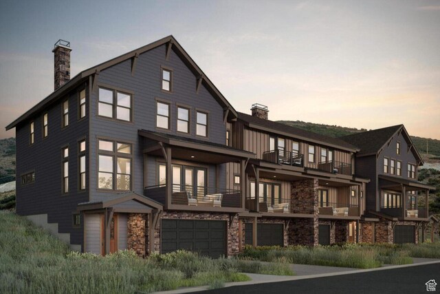 back of house at dusk with stone siding, a chimney, an attached garage, and a balcony