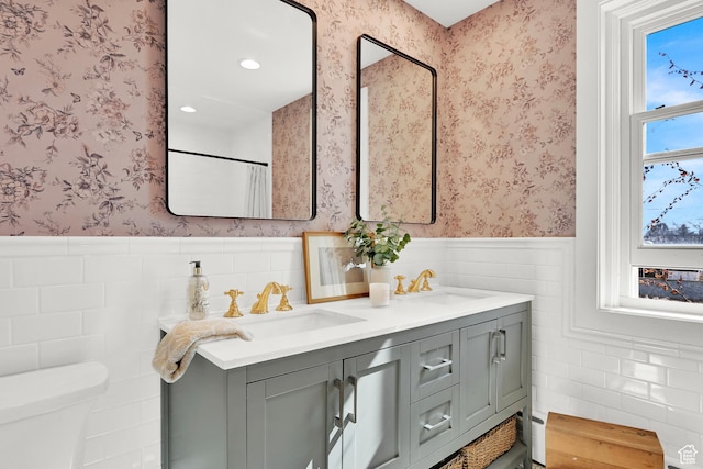 bathroom featuring a sink, toilet, double vanity, wainscoting, and wallpapered walls