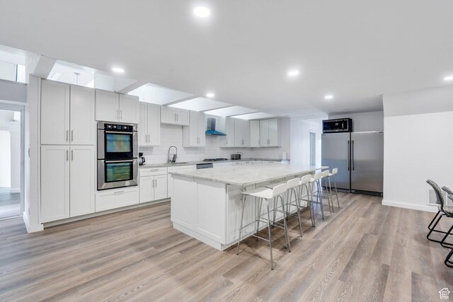 kitchen featuring stainless steel appliances, a sink, wall chimney range hood, light wood-type flooring, and decorative backsplash