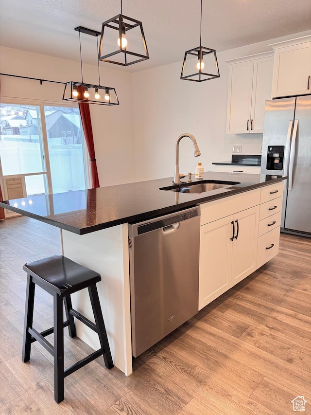 kitchen featuring light wood finished floors, an island with sink, a sink, stainless steel appliances, and dark countertops