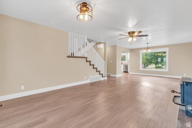 unfurnished living room with stairway, visible vents, baseboards, light wood-type flooring, and ceiling fan with notable chandelier