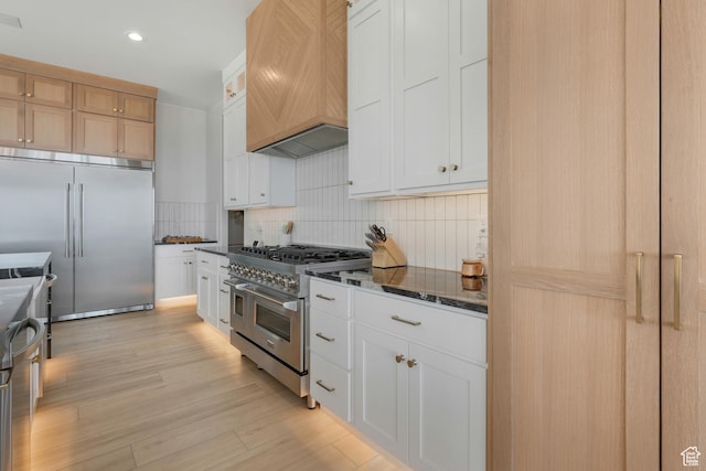 kitchen with premium range hood, dark stone counters, light wood finished floors, decorative backsplash, and high end appliances