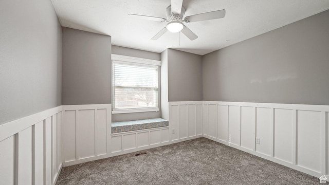 carpeted empty room with a decorative wall, wainscoting, ceiling fan, and visible vents