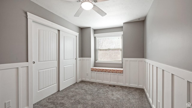 unfurnished bedroom featuring a wainscoted wall, carpet floors, visible vents, a ceiling fan, and a closet