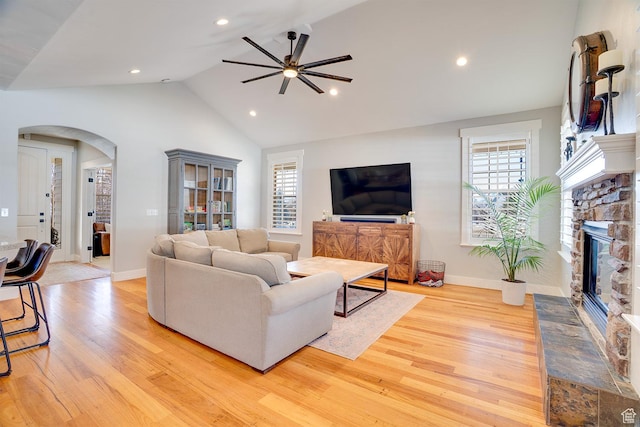 living room featuring light wood-style floors, ceiling fan, arched walkways, vaulted ceiling, and a stone fireplace