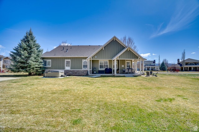 rear view of house with a hot tub, stone siding, a lawn, board and batten siding, and a patio