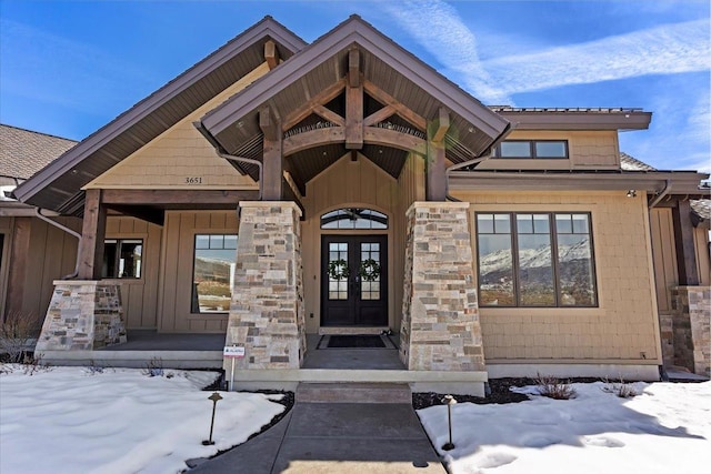 snow covered property entrance with french doors, stone siding, and board and batten siding