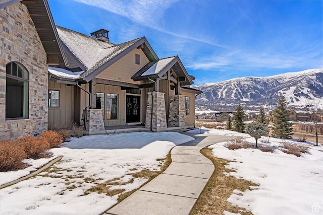 snow covered house with a mountain view, board and batten siding, stone siding, and a chimney