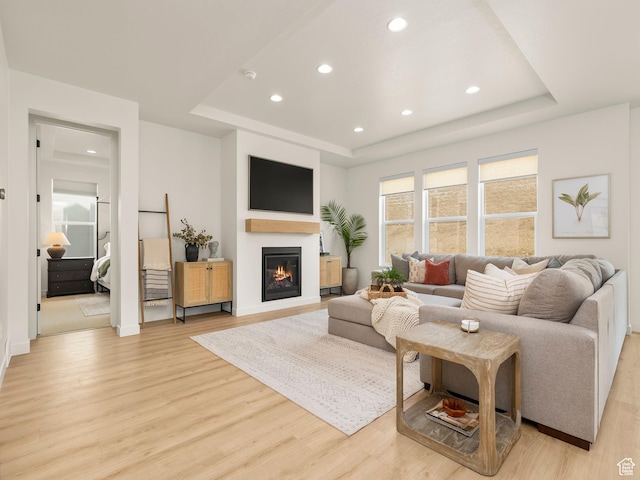 living room with a tray ceiling, a glass covered fireplace, recessed lighting, light wood-style floors, and baseboards