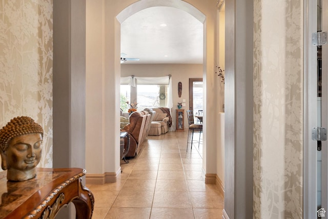hall featuring light tile patterned floors, baseboards, arched walkways, and wallpapered walls