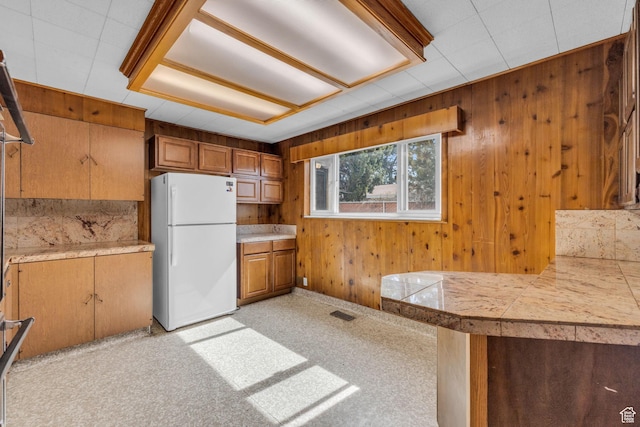 kitchen featuring tile countertops, brown cabinetry, visible vents, freestanding refrigerator, and wood walls