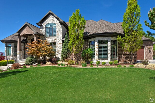 french country inspired facade with stone siding, a front lawn, a garage, a shingled roof, and brick siding