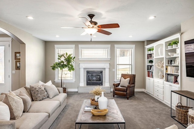 carpeted living room with arched walkways, a glass covered fireplace, plenty of natural light, and a ceiling fan