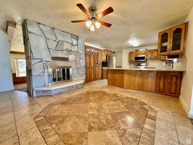 kitchen featuring brown cabinetry, a peninsula, stainless steel appliances, light countertops, and glass insert cabinets