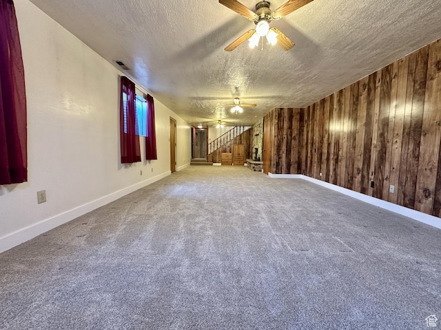 carpeted empty room with visible vents, a textured ceiling, stairway, wooden walls, and ceiling fan