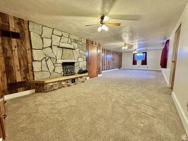 unfurnished living room with ceiling fan, wood walls, carpet flooring, a wood stove, and a textured ceiling