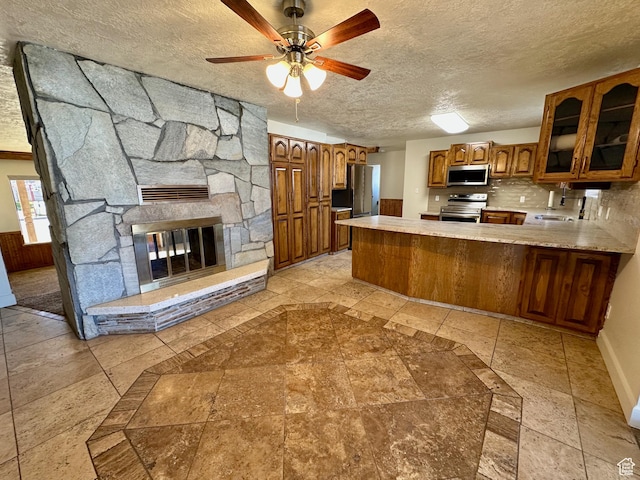 kitchen featuring backsplash, a textured ceiling, stainless steel appliances, brown cabinetry, and glass insert cabinets