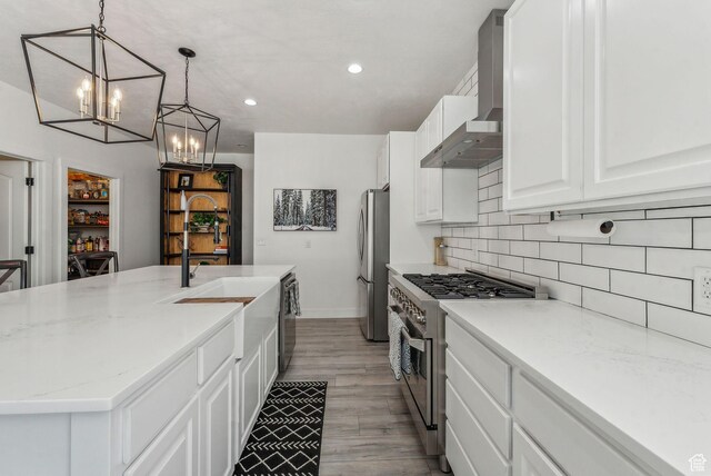 kitchen with a notable chandelier, a kitchen island with sink, a sink, stainless steel appliances, and wall chimney range hood