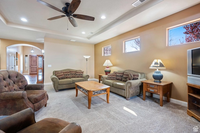 carpeted living room with a tray ceiling, arched walkways, visible vents, and ceiling fan