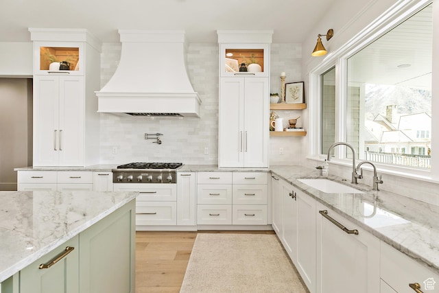 kitchen with stainless steel gas cooktop, custom range hood, light wood-style floors, white cabinetry, and a sink