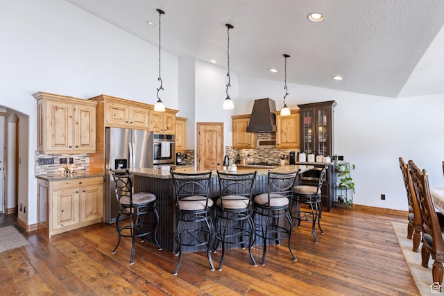 kitchen featuring dark wood finished floors, a breakfast bar, an island with sink, appliances with stainless steel finishes, and wall chimney exhaust hood